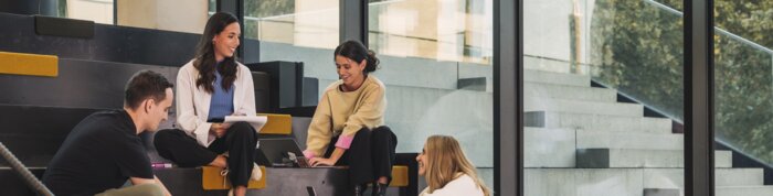 Studierende sitzen auf Treppen mit gelben und grünen Sitzkissen, lachen gemeinsam und lernen zusammen an einem Laptop. | © Lamprecht