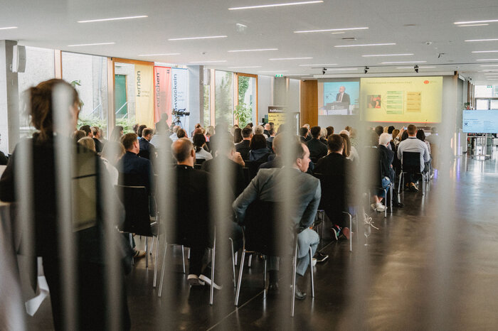 Publikum eines Business Summits, aufgenommen von hinten, während ein Redner auf der Bühne präsentiert. | © FHV