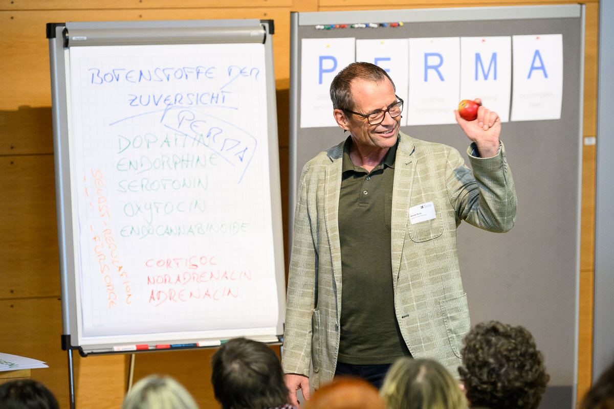 Referent steht vor einer Flipchart mit überschrift "Botenstoffe der Zuversicht" und hält einen Apfel in der Hand. | © Matthias Rhomberg | FHV