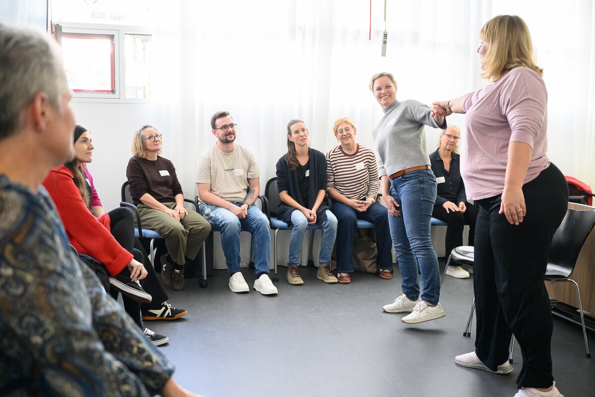 Menschen sitzen im Stuhlkreis. In der Mitte zwei Frauen, welche eine Vertrauensübung machen - sich halten und weit auseinander lehnen. | © Matthias Rhomberg | FHV
