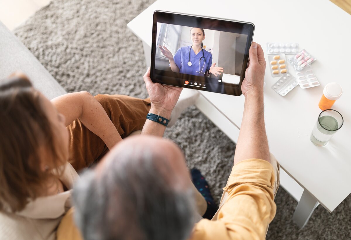 Zwei Menschen sitzen auf einer Bank und schauen gemeinsam auf ein Tablet, das der ältere Mensch in Händen hält. Auf dem Tablet-Bildschirm ist eine Pflegefachfrau zu sehen, die sich mit den Menschen unterhält.