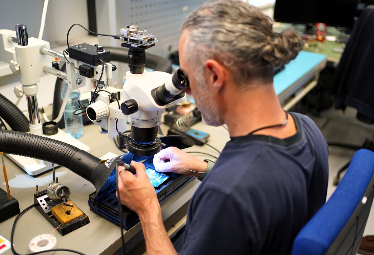 Ein Mann in einem Blaumann sitzt, schaut durch ein Mikroskop und lötet gleichzeitig. | © Enter Electronics
