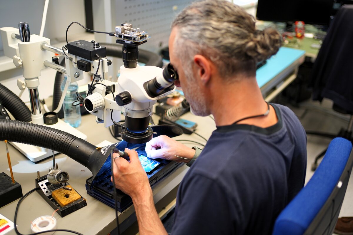 Ein Mann in einem Blaumann sitzt, schaut durch ein Mikroskop und lötet gleichzeitig. | © Enter Electronics