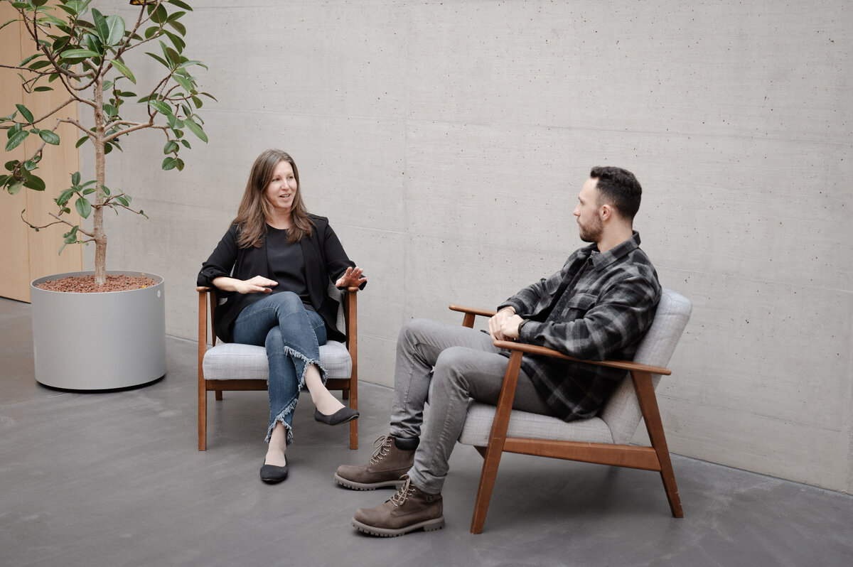 Sabrina und Markus sitzen auf Sesseln vor einer grauen Betonwand und unterhalten sich miteinander. | © FHV