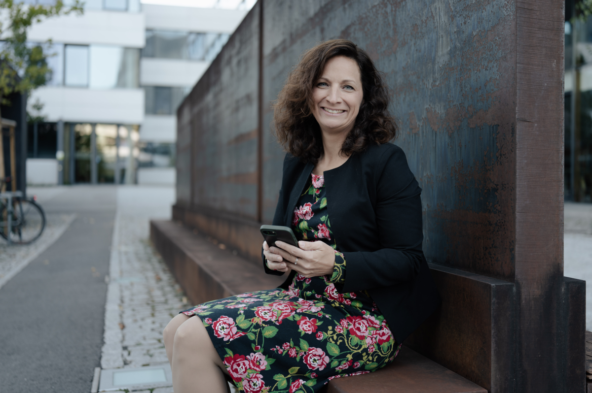 Wissenschaftlerin Sarah Kühne sitzt lächelnd mit ihrem Handy in der Hand auf einer Bank. | © FHV