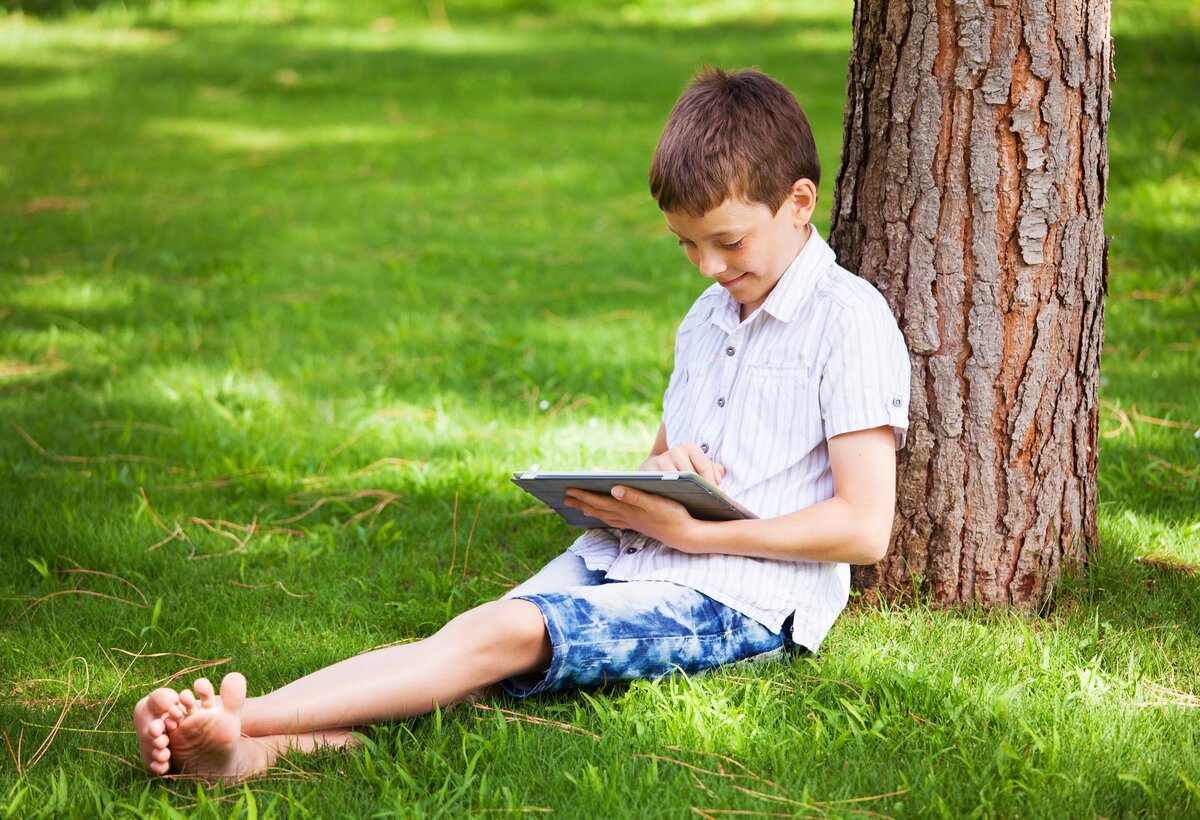 Ein Junge sitzt im Gras und lehnt an einem Baum. Er hat ein Tablet in der Hand.