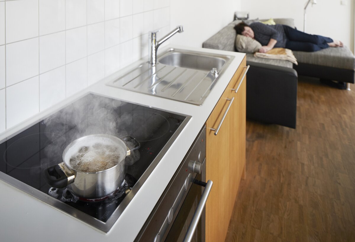 Kochtopf steht auf eingeschaltenem Herd und ältere Person liegt auf dem Sofa.