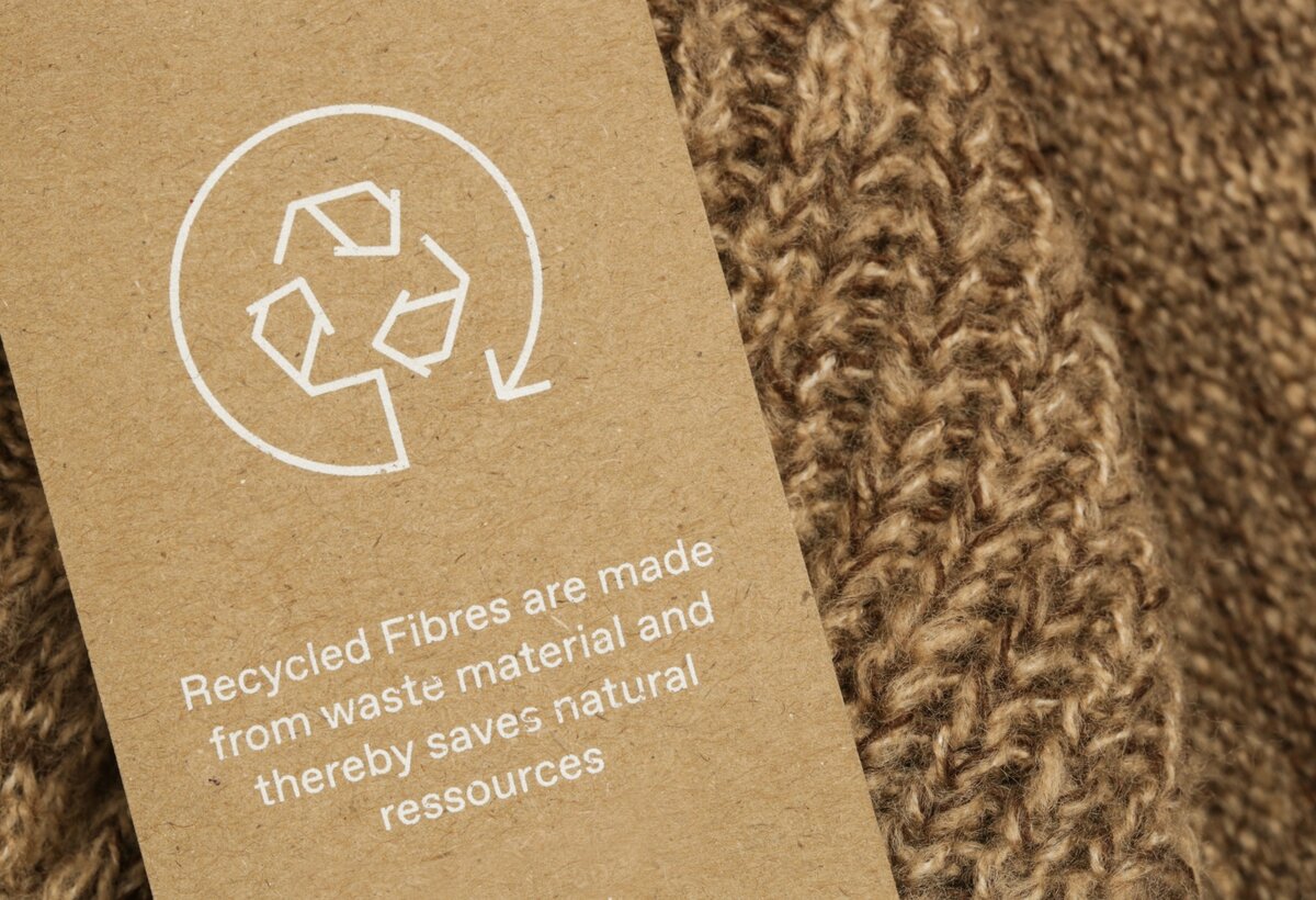 Brauner Strick mit einem Etikett aus Recyclingpapier, das auf recycelte Fasern und Ressourcenschonung hinweist. | © Adobe Stock