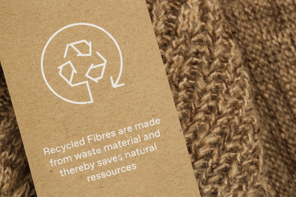 Brauner Strick mit einem Etikett aus Recyclingpapier, das auf recycelte Fasern und Ressourcenschonung hinweist. | © Adobe Stock