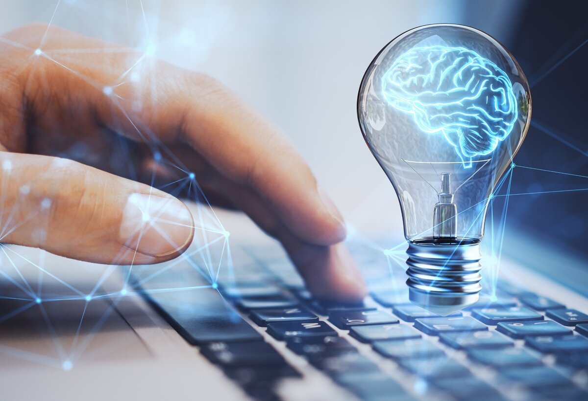 Hand über Tastatur, darüber schwebt eine leuchtende Glühbirne mit blauem Gehirn als Symbol für kreative Ideen. | © Adobe Stock