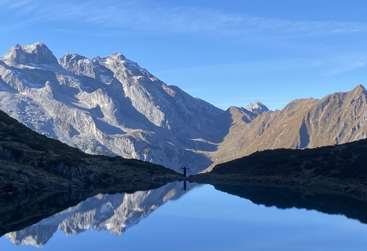 Alpine Scenery with a mirror lake and mountains, in the distance a silouette of aperson spreading their arms. | © FHV