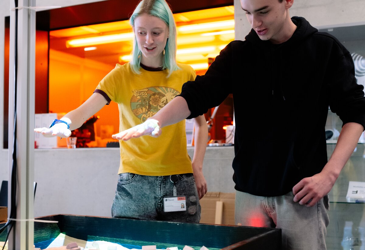 Fabrizia (links, BORG Schoren) und Maximilian (rechts, HTL Dornbirn) stehen vor der Augmented Reality-Sandbox und lassen es mit ihren Händen regnen. | © FHV Zwei Schüler:innen stehen vor der Augmented Reality-Sandbox und lassen es mit ihren Händen regnen | © FHV