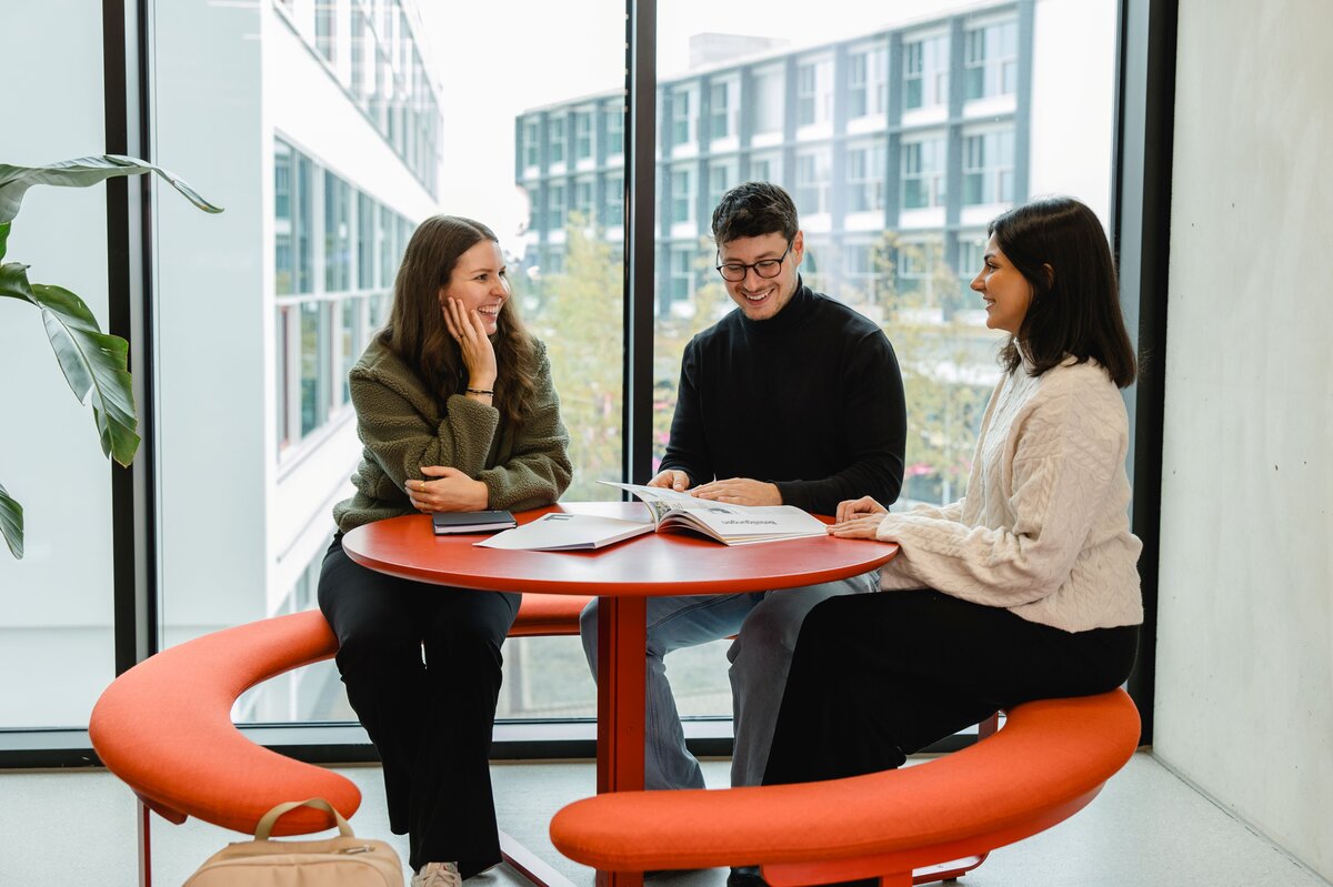 Lachende Studierende sitzen auf einer roten Sitzbank an einem Tisch und unterhalten sich über den Studienalltag. Im Hintergrund ist der FHV-Campus zu sehen. | © FHV | Broell