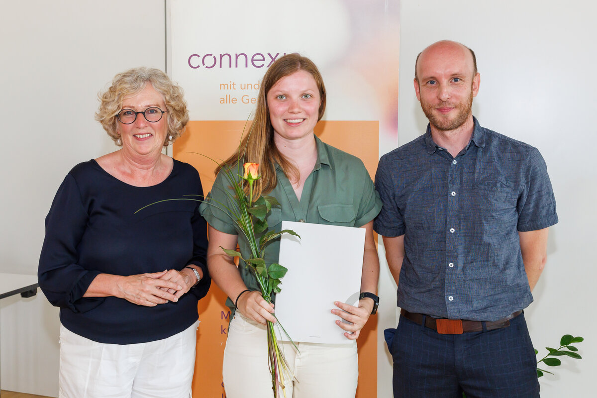 Studentin steht vor einem Roll-up-Banner der Connexia, hält ihr Zertifikat zur Praxisanleiterin-Ausbildung und eine Rose. Neben ihr steht links eine Frau und rechts ein Mann. | © Privat