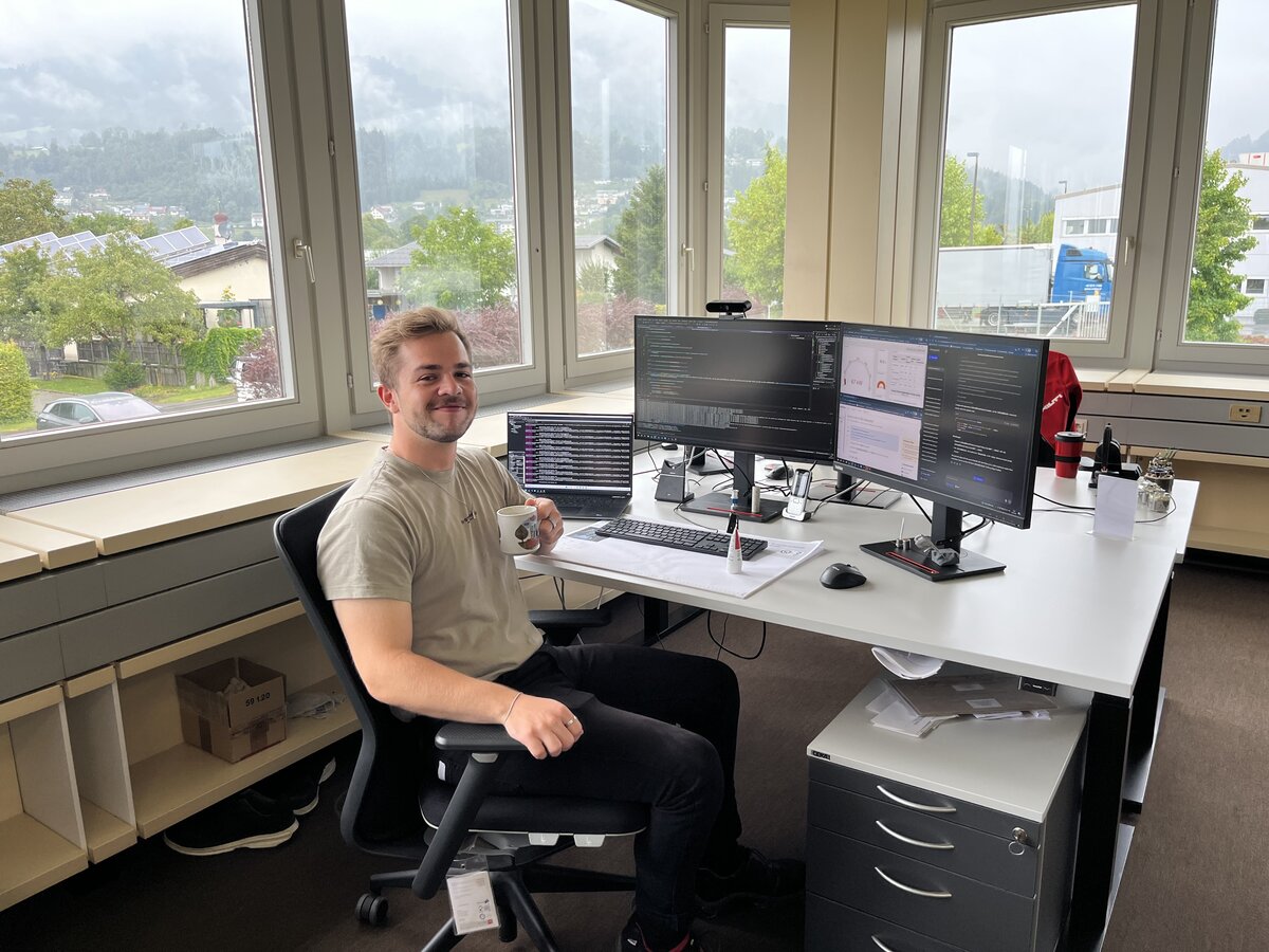 Jonathan sitzt an seinem Arbeitsplatz mit einer Tasse Kaffee. Durch das Fenster im Hintergrund ist ein schönes Bergpanorama zu sehen. | © FHV