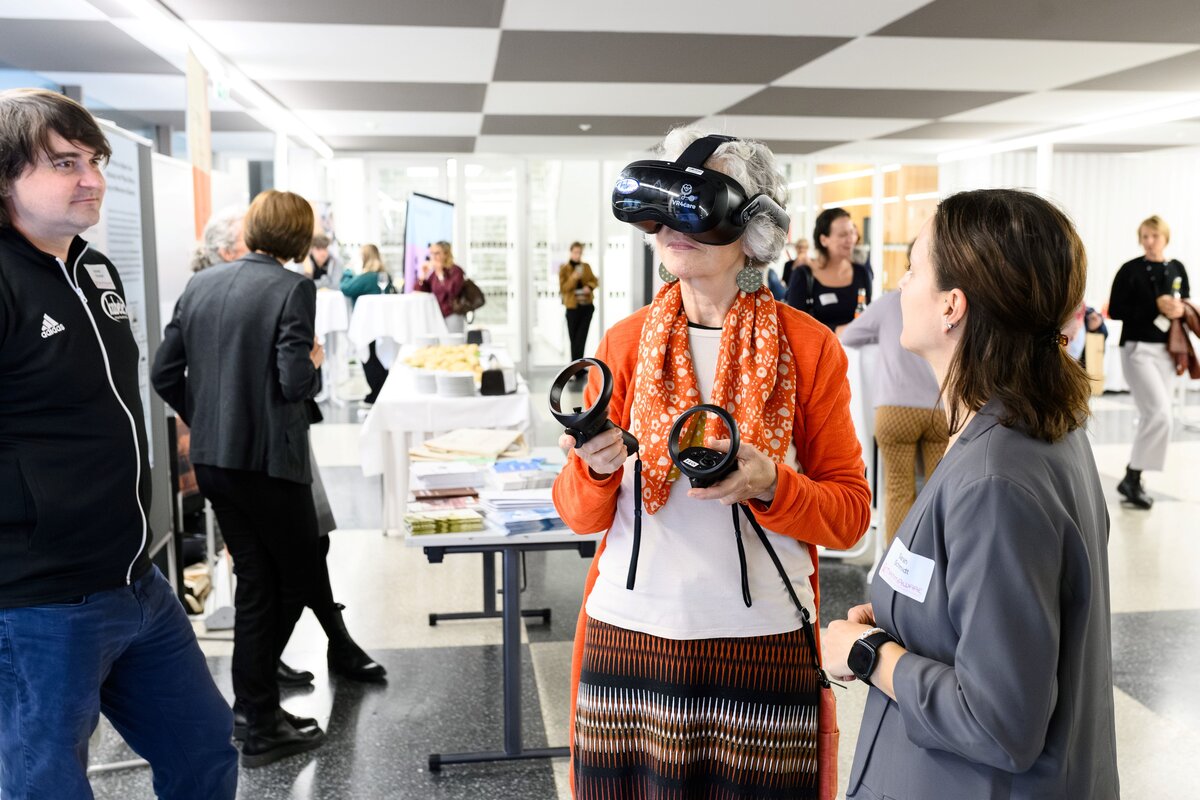 Dame hat einen VR-Brille auf und wird von zwei Kollegen der FHV betreut. Im Hintergund stehen einige andere Menschen und unterhalten sich. | © Rhomberg | FHV