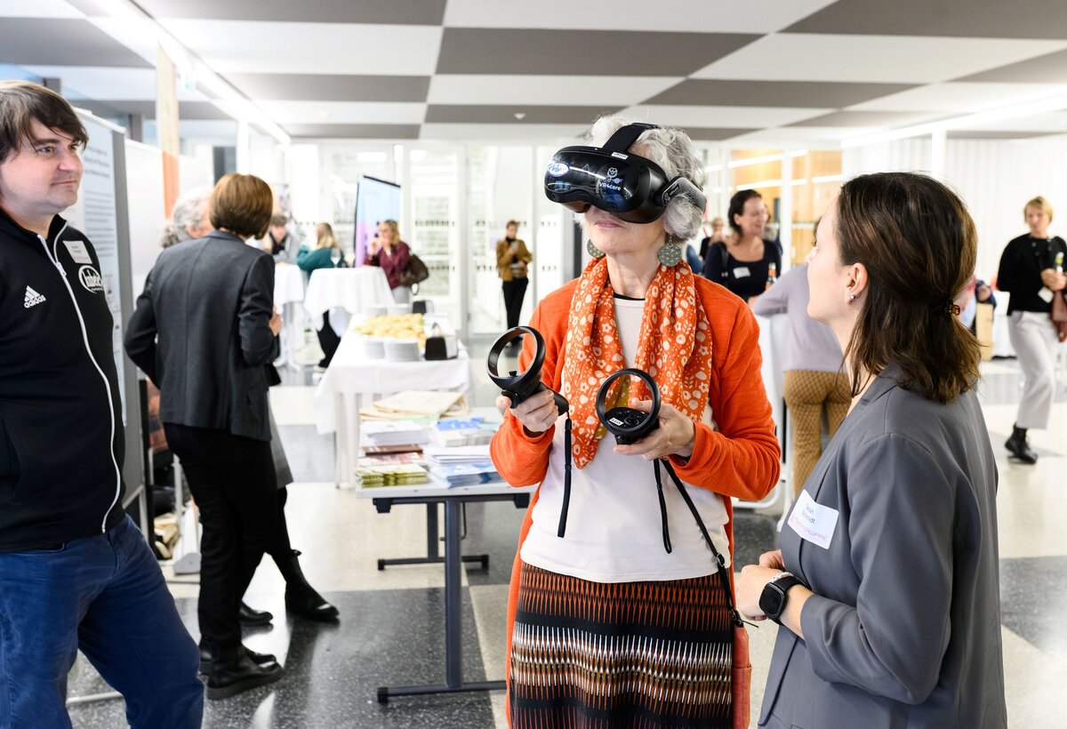 Dame hat einen VR-Brille auf und wird von zwei Kollegen der FHV betreut. Im Hintergund stehen einige andere Menschen und unterhalten sich. | © Rhomberg | FHV