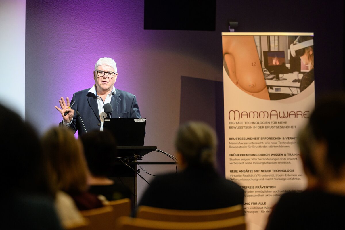 Dr. Hubert Jocham steht auf der Bühne, umgeben von violettem Licht. Rechts von ihm befindet sich ein Plakat mit dem Schriftzug ‚MammAware‘. Im Vordergrund sitzt das Publikum von hinten auf Stühlen. | © Rhomberg | FHV
