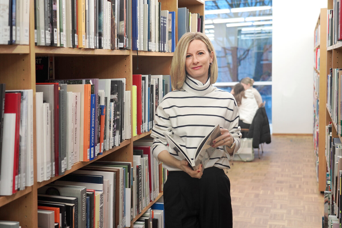 Eine Frau hält ein aufgeschlagenes Buch in den Händen, steht in einer Bibliothek und lächelt in die Kamera. | © FHV