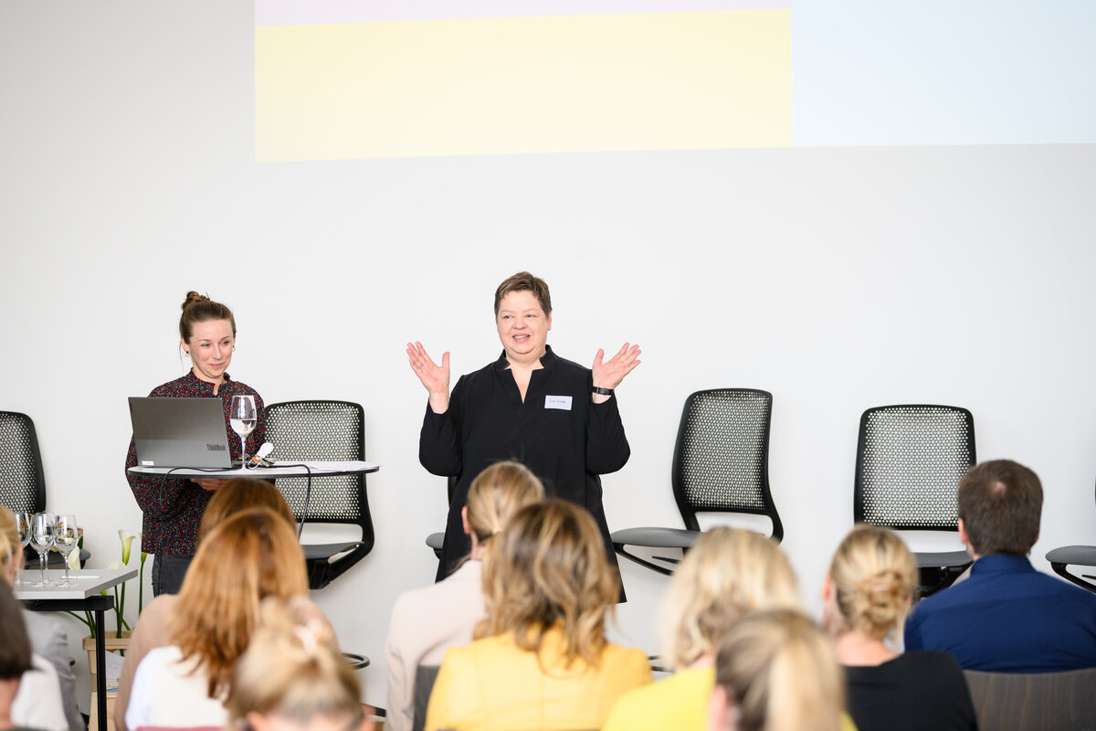 Diana Brodda steht auf der Bühne eines Pflegesymposiums und hält einen Vortrag. Sie spricht engagiert zum Publikum, während hinter ihr eine Präsentationsfolie zu sehen ist. Die Atmosphäre wirkt fachlich konzentriert, die Zuhörer verfolgen aufmerksam ihre Ausführungen. | © Rhomberg | FHV