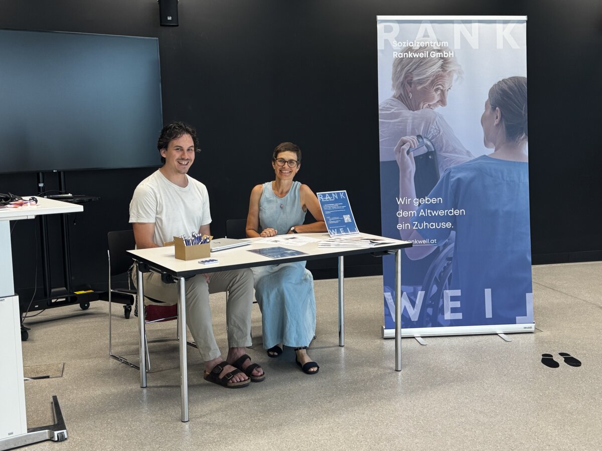 Zwei Ausstellende sitzen an einem Tisch und neben ihnen ist ein Rollup-Banner des Sozialzentrum Rankweil. | © FHV
