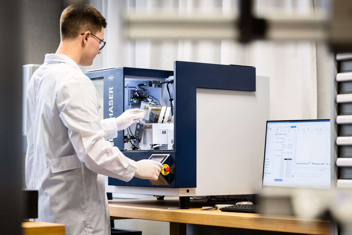 Ein Mann mit Handschuhen und weißem Laborkittel steht an einer Maschine. Neben ihm werden auf einem PC Daten angezeigt. | © FHV | Rhomberg