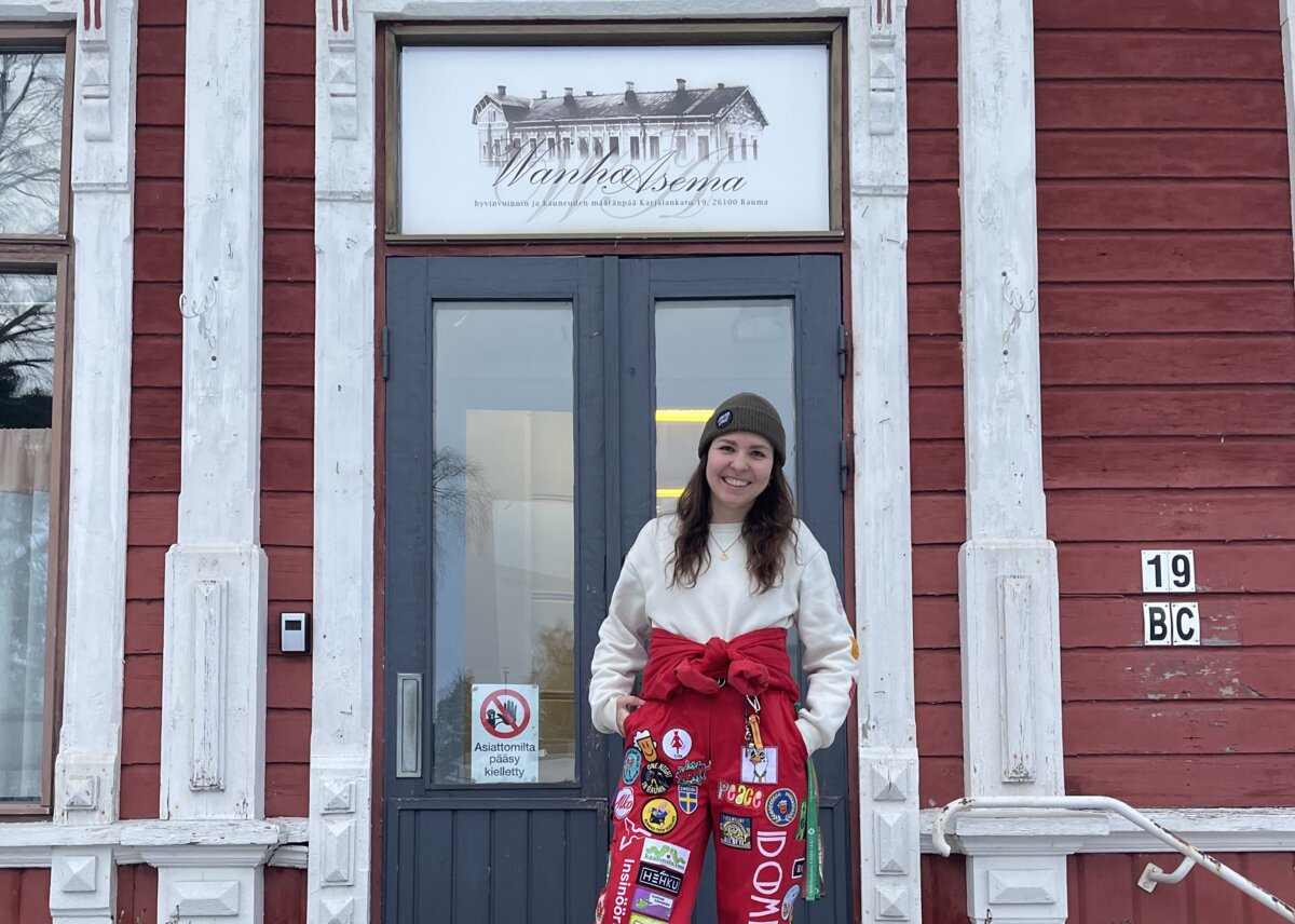Valentina Angeli steht vor dem roten Holzgebäude der Old Station, ihrer Studierendenunterkunft im finnischen Rauma. Auf den Treppen liegt Schnee, sie trägt einen roten Overall mit bunten Patches. | © privat