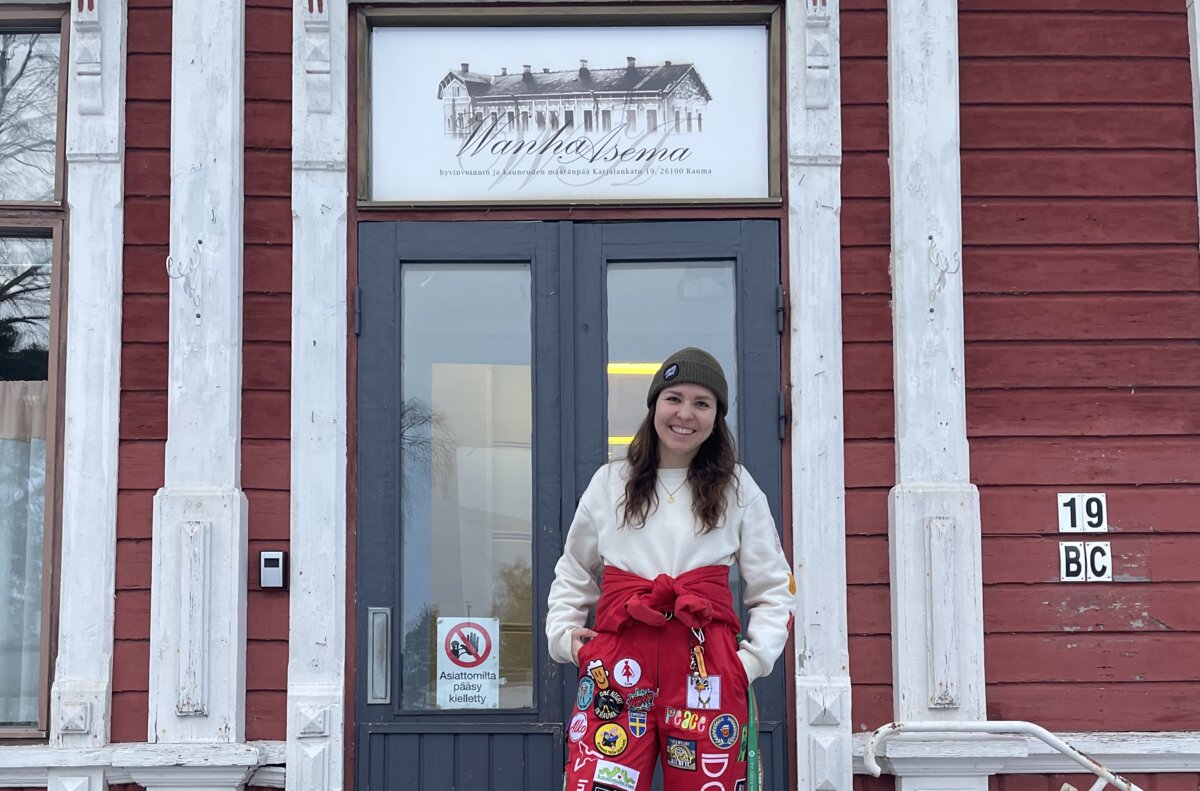Valentina Angeli steht vor dem roten Holzgebäude der Old Station, ihrer Studierendenunterkunft im finnischen Rauma. Auf den Treppen liegt Schnee, sie trägt einen roten Overall mit bunten Patches. | © privat