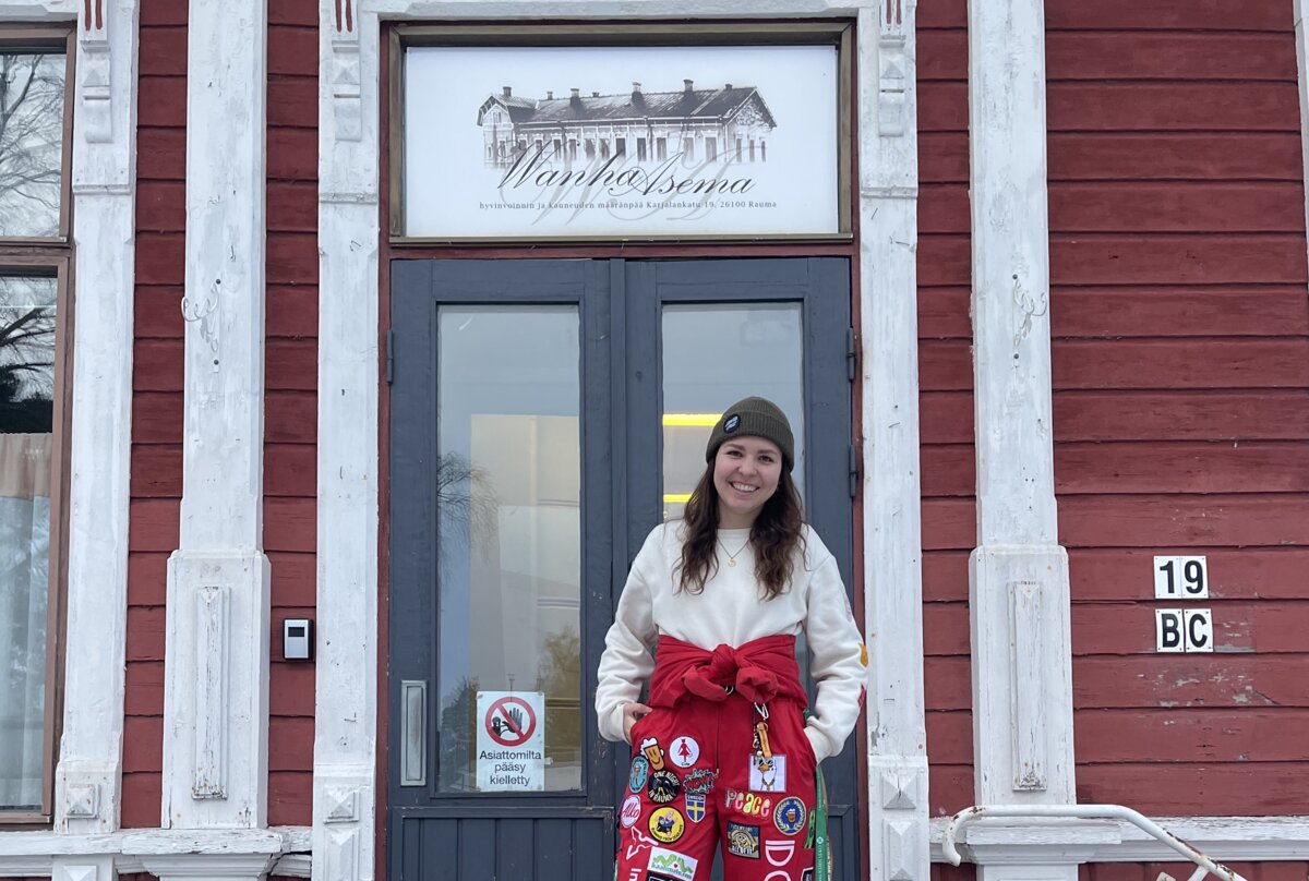 Valentina Angeli steht vor dem roten Holzgebäude der Old Station, ihrer Studierendenunterkunft im finnischen Rauma. Auf den Treppen liegt Schnee, sie trägt einen roten Overall mit bunten Patches. | © privat